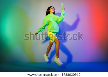 Full length body size view of attractive cheerful girl jumping using device isolated over multicolor vivid neon light background Royalty-Free Stock Photo #1988030912