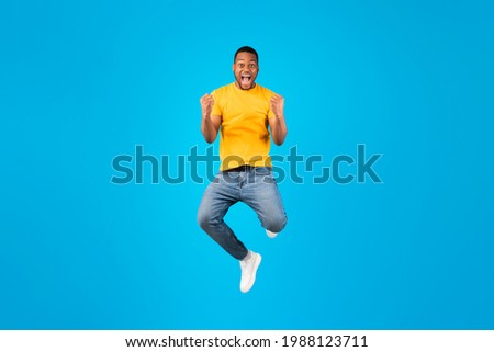 Joy Of Victory. Emotional Black Guy Gesturing Yes And Shouting Posing In Mid-Air Over Blue Background. Millennial Man Jumping And Shaking Fists Looking At Camera Celebrating Success In Studio