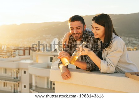 Couple enjoying cityscape and seaside view, drinking beer and eating pizza on the rooftop. Portrait of happy people spend quarantine time, picnic leisure on the roof on the sunset.