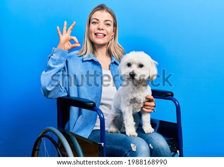 Beautiful caucasian woman sitting on wheelchair hugging dog doing ok sign with fingers, smiling friendly gesturing excellent symbol 