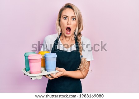 Beautiful young blonde woman wearing waitress apron holding take away cup of coffees afraid and shocked with surprise and amazed expression, fear and excited face. 