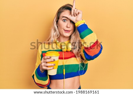 Beautiful young blonde woman drinking cup of coffee wearing headphones making fun of people with fingers on forehead doing loser gesture mocking and insulting. 