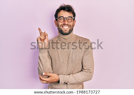 Handsome man with beard wearing turtleneck sweater and glasses with a big smile on face, pointing with hand and finger to the side looking at the camera. 