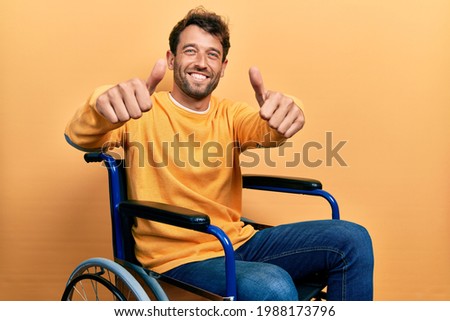 Handsome man with beard sitting on wheelchair approving doing positive gesture with hand, thumbs up smiling and happy for success. winner gesture. 