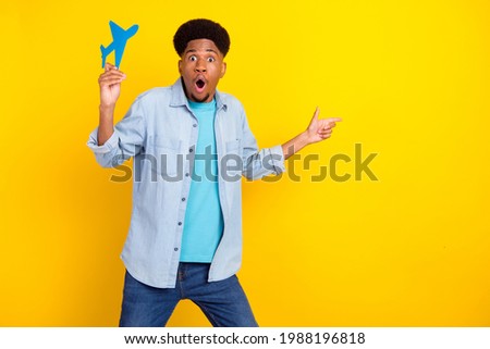 Photo of funny shocked dark skin man wear jeans shirt holding paper plane pointing empty space isolated yellow color background