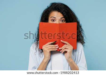 Young shocked puzzled nerd african american girl teen student in denim clothes cover mouth hiding with book notebook isolated on blue background Education in high school university college concept.