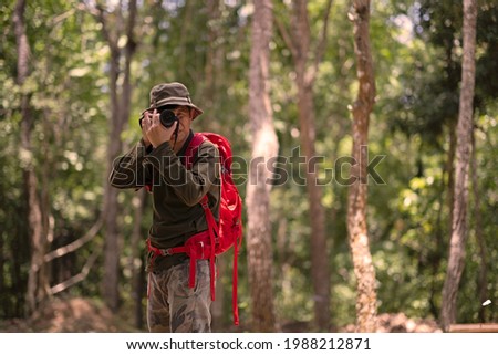An asian senior photographer using digital camera to took the photos, a man in green long sleeve t-shirt and camouflage pants and hat pose taking photo action to camera with forest background.