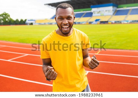 Emotional young afro american sportsman iin yellow trendy t-shirt celebrating victory, goal, success making winner gesture with clenched fists, screaming on the stadium