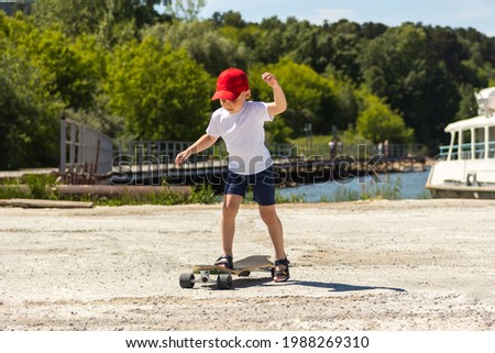 A little boy in a white t-shirt, red cap, shorts and sandals rides a skateboard on the background of the pier. A sporty caucasian kid walking around. Children's leisure. Wellness