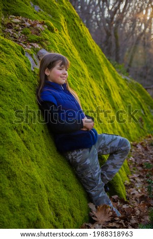 A young girl with long brown hair in a sleeveless jacket near a stone with green moss as a wall in the forest in early spring against a background of sunset