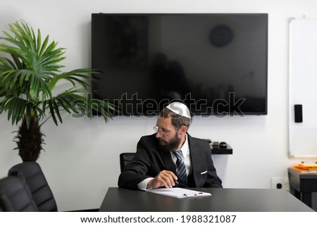 Jewish man in a yarmulke and glasses writing down notes in clipboard while working in office. Jew man in a white shirt, business suit and national hat Kippah and and looking to the side his brows bent