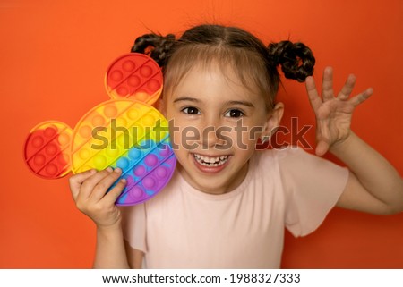 An emotional portrait of a cute little girl on an orange background with a funny hairstyle made of pigtails and a popit toy  in her hands. Hand gestures. Pop it toy