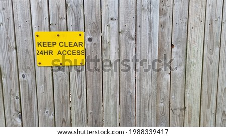 A wooden fence with planks running from top to bottom with a yellow plastic sign in the left upper corner displaying the words keep clear 24hr access in black text with copy space to the right.