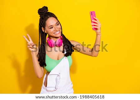 Photo of brunette dark skin young woman wear green top white overall make selfie v-sign isolated on yellow color background
