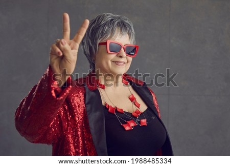Happy grandma in red and black sequin party outfit and cool glasses smiling and doing victory gesture. Funny retired old senior woman showing peace sign standing on gray studio background