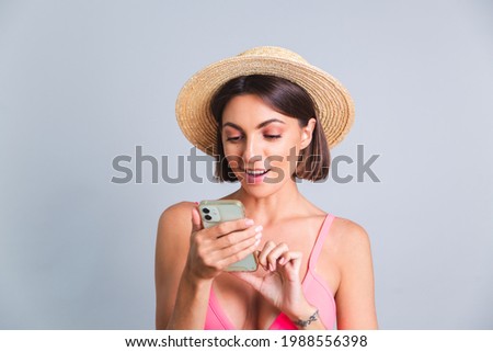 Pretty woman in bikini and straw hat on gray background positive thoughtful typing message on mobile phone