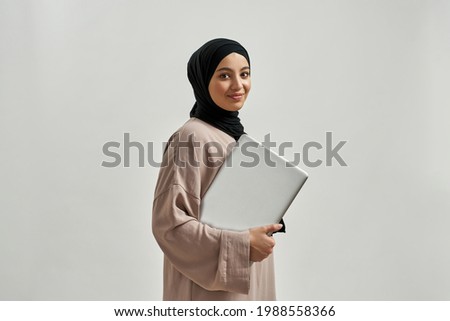 Happy young arabian girl holding laptop while posing on light background and smiling at camera. Beautiful muslim lady Royalty-Free Stock Photo #1988558366