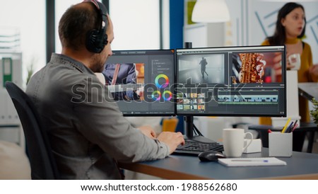 Videographer putting on headset editing video project using post production software working in in digital multimedia company with two monitors. Retoucher processing audio film montage on computer