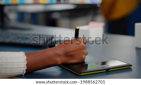 Close up on hand of african photo editor drawing on graphic tablet editing customer photo on computer with two monitors. Black retoucher holding stylus pen retouching assets in creative agency office