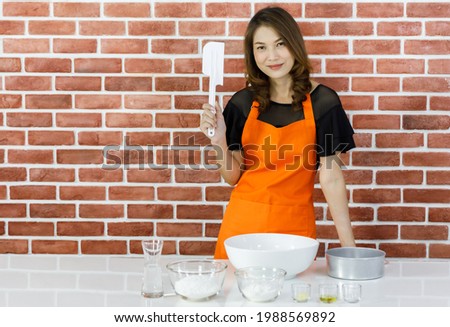 Lovely young Asian woman in orange apron smilingly standing near brick wall and holding spatula and metal sieve as ready to enjoy happy cooking with kitchenwares on white table in home kitchen