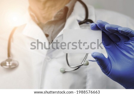 Old doctor wearing protective medical glove holds blank white card with mockup space for advertise information