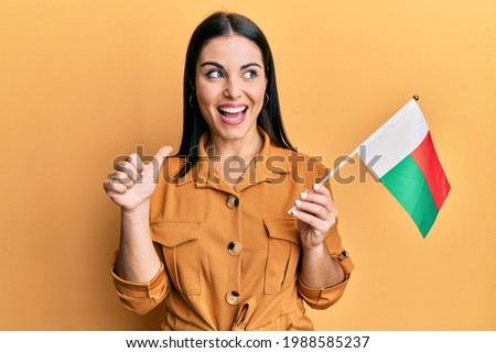 Young brunette woman holding madagascar flag pointing thumb up to the side smiling happy with open mouth 