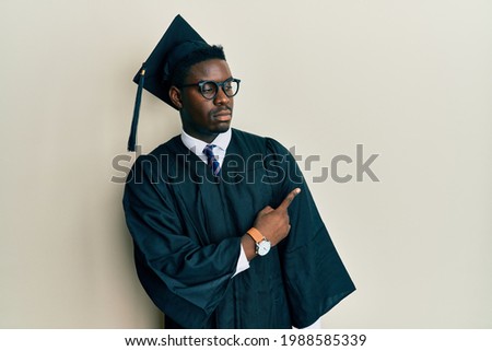 Handsome black man wearing graduation cap and ceremony robe pointing with hand finger to the side showing advertisement, serious and calm face 