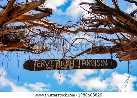 "Welcome Back" signage in english and finnish language hanged in between dead trees against a blue and partially cloudy sky.