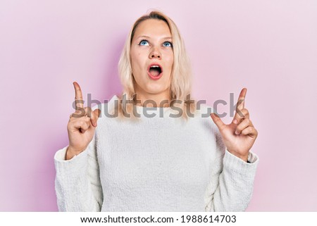 Beautiful caucasian blonde woman wearing casual winter sweater amazed and surprised looking up and pointing with fingers and raised arms. 