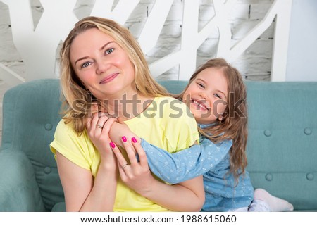 Little caucasian girl sitting together with mother on the sofa in living room and hugging each other smiling at the camera. Spending time with children at home.