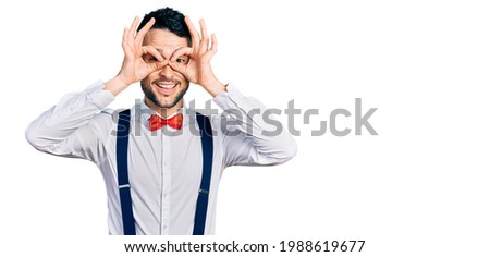 Hispanic man with beard wearing hipster look with bow tie and suspenders doing ok gesture like binoculars sticking tongue out, eyes looking through fingers. crazy expression. 