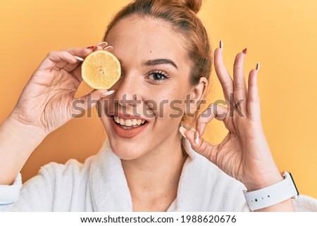Young blonde woman wearing bathrobe holding lemon over eye doing ok sign with fingers, smiling friendly gesturing excellent symbol 