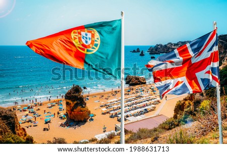 Portugal and United Kingdom flags on foreground with perspective view of beachgoers at Cova Redonda Beach in Algarve, southern Portugal on a summer day