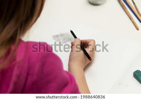 little girl sits at a white table, holds a pencil in her hand and is engaged in drawing, back view, lifestyle