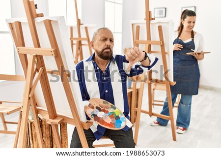 Senior artist man at art studio looking unhappy and angry showing rejection and negative with thumbs down gesture. bad expression. 