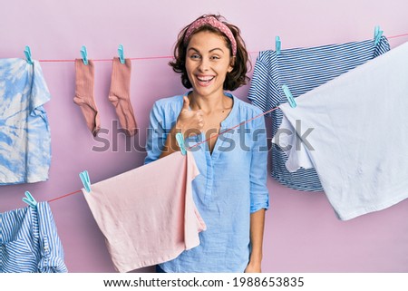 Young brunette woman doing laundry around string hangs doing happy thumbs up gesture with hand. approving expression looking at the camera showing success. 