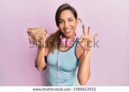 Young latin woman wearing gym clothes, using headphones and holding cornflakes doing ok sign with fingers, smiling friendly gesturing excellent symbol 