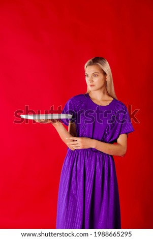 Smiling blondie in purple dress holding mockup plate. Attractive blonde girl model in long summer dress holding a white mockup plate on red matte background. Presenting and advertising concept.