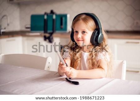 Smiling little girl sitting in the kitchen at home, watching video on smartphone, happy child in headphones, having fun, talking. kids and gadgets, blogger, online party, streaming. technology concept