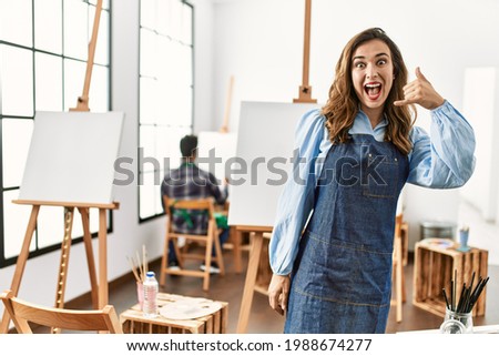 Young artist woman at art studio smiling doing phone gesture with hand and fingers like talking on the telephone. communicating concepts. 