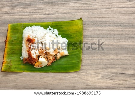 A picture of "Nasi Lemak Seringgit' on copyspace woodedn table. Coconut rice serve with sauce, nuts and anchovies wrap in banana leaf that cost one 1 Ringgit.