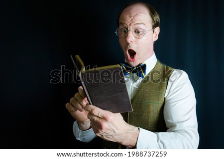 Portrait of Nerdish Fellow in Vest and Bow Tie Reading a Book with an Expression of Amazement on Black Background. 