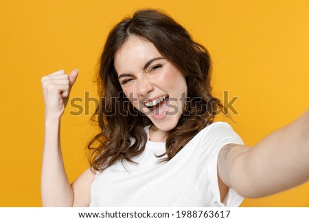 Close up young excited overjoyed happy cool caucasian woman 20s in white basic casual t-shirt do selfie shot on mobile phone do winner gesture clench fist isolated on yellow background studio portrait