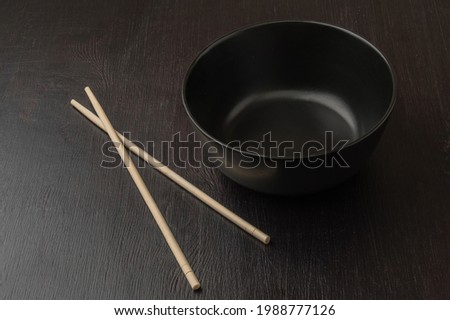 The wooden chopsticks and black ceramic bowl are on the black wooden table. Dark food photography of empty plate and sticks.