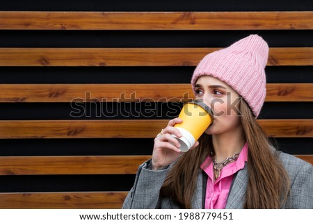 A 22-year-old girl in fashionable clothes drinks coffee from a yellow plastic cup, dressed in a gray coat, a pink shirt and a pink hat on a background of wooden boards. Copy space. High quality photo