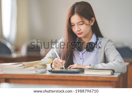 Stock photo of a young teenage woman Asian college student in student uniform studying and writing on digital tablet in a university classroom