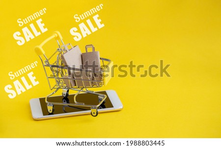 Metal cart with paper bags for goods, stands on cell phone. Text-summer sale is written on yellow isolated background. Concept of summer online shopping.