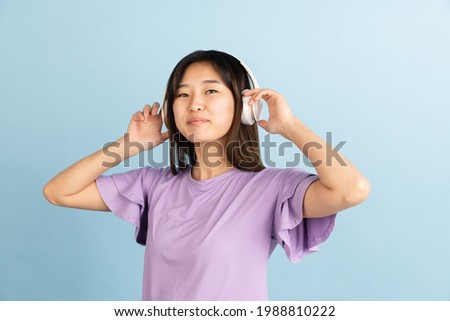 Listening to music with headphones. Asian young woman's portrait on blue studio background. Beautiful female model in casual style. Concept of human emotions, facial expression, youth, sales, ad.