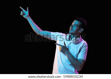 Pointing, choosing. Latina young man's portrait isolated on dark studio background in neon light. Handsome male model in headphones. Concept of human emotions, facial expression, youth, sales, ad.