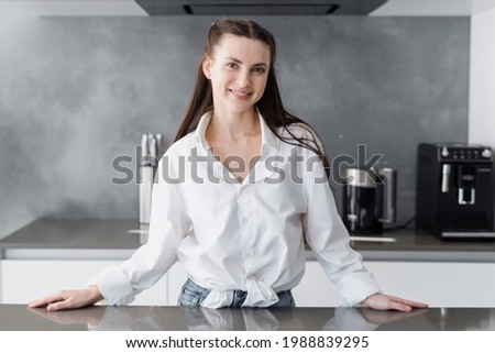 Young happy woman standing behind countertop, preparing to cooking alone in modern kitchen interior, looking at camera, smiling wide. Charming housewife in casual clothes spending morning at home Royalty-Free Stock Photo #1988839295